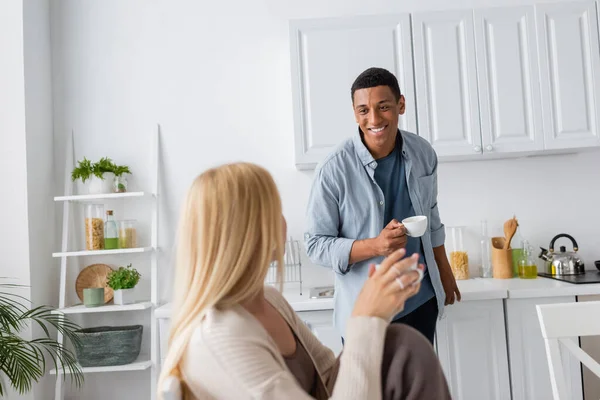 Happy african american man with coffee cup looking at blonde girlfriend on blurred foreground — Stock Photo