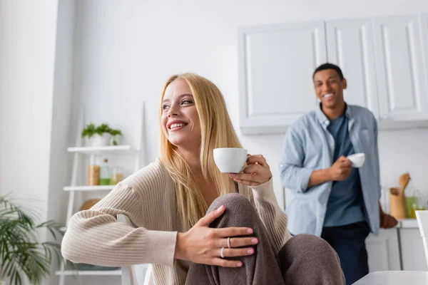 Mulher loira com xícara de café sorrindo e olhando para longe perto de namorado americano africano borrado — Fotografia de Stock