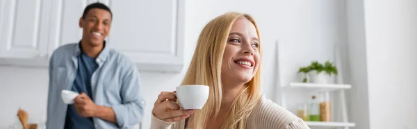 Femme blonde tenant tasse de café et regardant loin près de l'homme afro-américain sur fond flou, bannière — Photo de stock