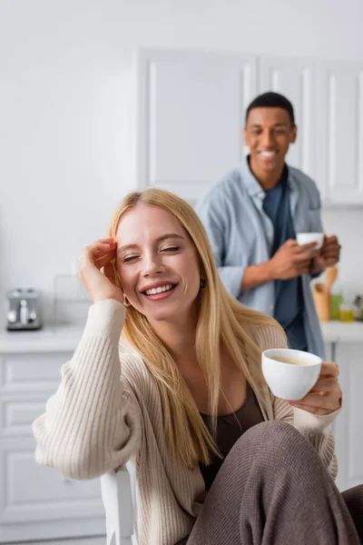 Mujer joven y feliz con los ojos cerrados sosteniendo taza de café cerca de novio afroamericano sonriendo sobre fondo borroso - foto de stock