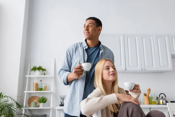 Africano americano uomo con caffè tazza guardando lontano vicino sorridente bionda fidanzata seduta con gli occhi chiusi in cucina — Foto stock