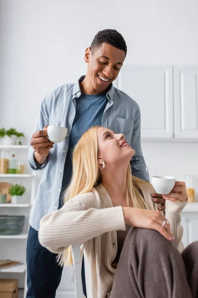 Cheerful interracial couple holding morning coffee and smiling at each other in kitchen — Stock Photo
