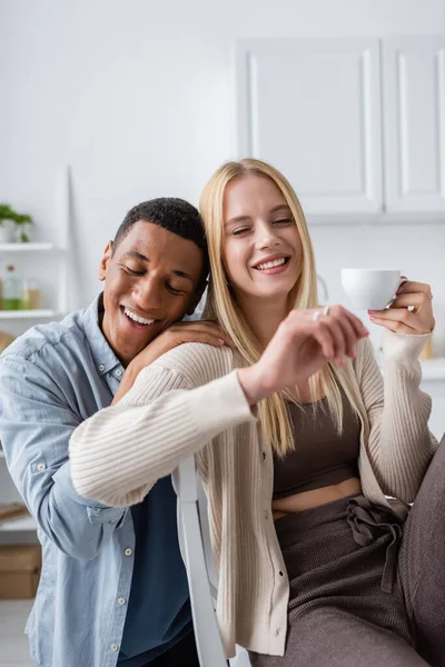 Homem americano africano satisfeito com olhos fechados abraçando namorada loira segurando xícara de café — Fotografia de Stock