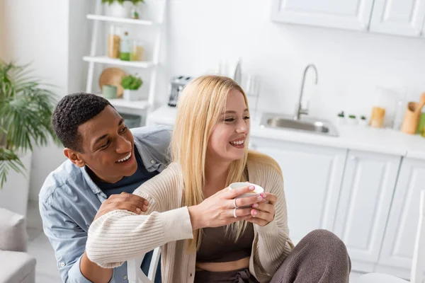 Afro-américain homme souriant près de jeune copine blonde assis avec café du matin dans la cuisine — Photo de stock