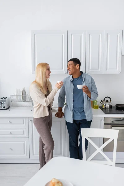 Glückliches interrassisches Paar trinkt Morgenkaffee und redet in moderner Küche — Stockfoto