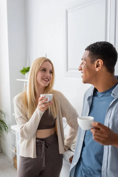 Young blonde woman and african american man smiling at each other while holding coffee cups in kitchen — Stock Photo