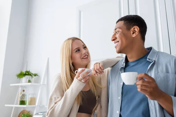 Alegre pareja interracial con tazas de café mirándose en la cocina - foto de stock