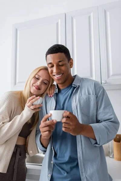 Улыбающаяся многонациональная пара, держащая утренний кофе на кухне — стоковое фото