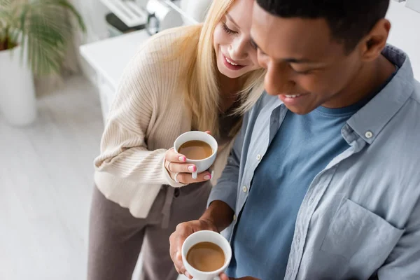 Vista ad alto angolo di felice coppia multietnica in piedi con tazze di caffè in cucina — Foto stock