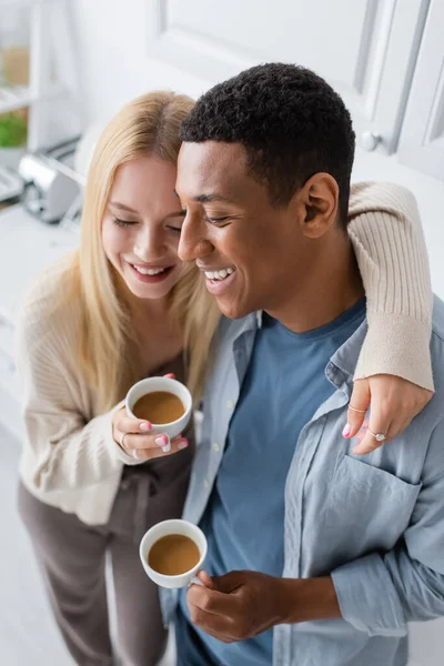 Hochwinkelaufnahme einer blonden Frau mit geschlossenen Augen und einem glücklichen afrikanisch-amerikanischen Mann, der mit Kaffeetassen in der Küche steht — Stockfoto