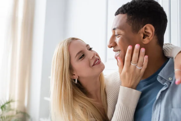 Happy blonde woman hugging african american boyfriend and touching his face in kitchen — Stock Photo