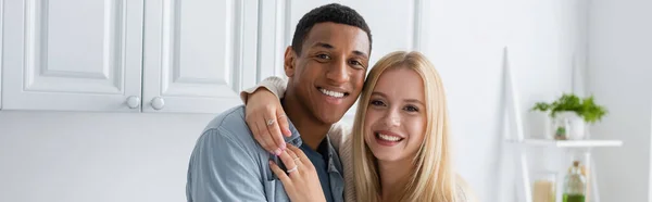 Young blonde woman hugging happy african american boyfriend while smiling at camera in kitchen, banner — Stock Photo