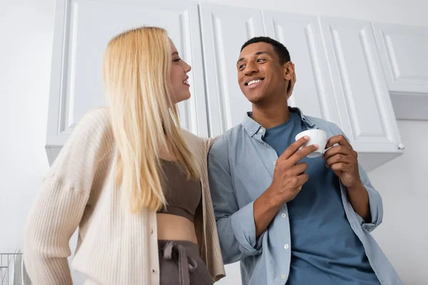 Low angle view of young blonde woman and african american man with coffee cup smiling at each other in kitchen — Stock Photo