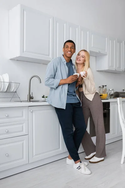 Piena lunghezza di felice coppia interrazziale in piedi con tazze di caffè in cucina moderna — Foto stock