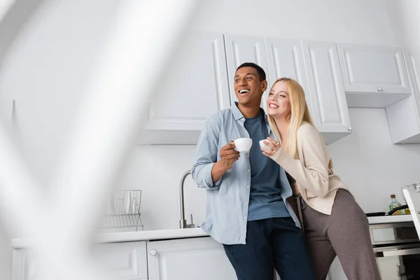 Возбужденная межрасовая пара с кофейными чашками глядя в сторону на кухне на размытом переднем плане — стоковое фото
