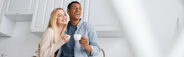Alegre pareja multiétnica con café de la mañana mirando hacia otro lado en primer plano borrosa en la cocina, pancarta - foto de stock