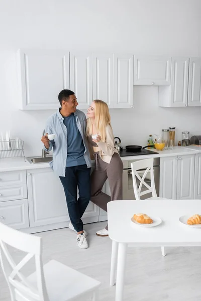 Alegre pareja interracial con tazas de café mirándose unos a otros cerca de croissants en la mesa en la cocina espaciosa y moderna - foto de stock
