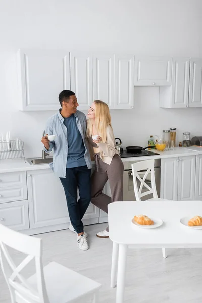 Freudiges gemischtrassiges Paar mit Kaffeetassen, die sich bei Croissants auf dem Tisch in der geräumigen und modernen Küche anschauen — Stockfoto