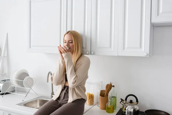 Mujer rubia con los ojos cerrados sentado en la encimera de la cocina y disfrutando del sabor del café de la mañana - foto de stock