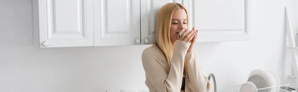 Молодая блондинка с закрытыми глазами пьет утренний кофе на кухне, баннер — стоковое фото