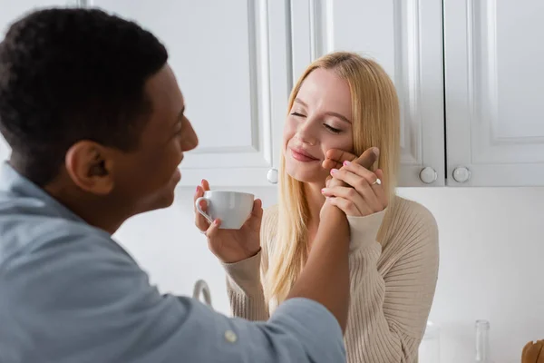 Femme blonde souriante avec les yeux fermés tenant tasse de café près floue homme afro-américain toucher son visage dans la cuisine — Photo de stock