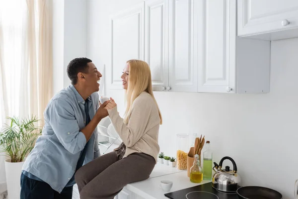 Vista lateral da mulher rindo sentado na bancada da cozinha e de mãos dadas com o namorado americano africano feliz — Fotografia de Stock