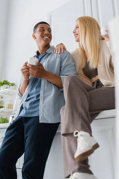 Baixo ângulo vista de casal interracial com xícaras de café sorrindo um para o outro na cozinha — Fotografia de Stock