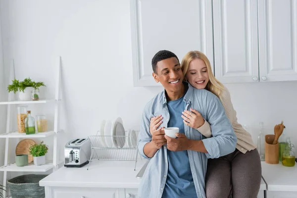 Joyful blonde woman with closed eyes sitting on kitchen worktop and hugging african american boyfriend standing with coffee cup — Stock Photo