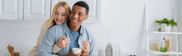 Fröhlicher afrikanisch-amerikanischer Mann mit Kaffeetasse, während junge Freundin ihn in der Küche umarmt, Banner — Stockfoto