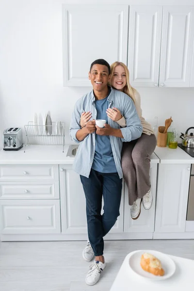Счастливая блондинка, сидящая на кухонном столе и обнимающая африканского американского парня, держащего чашку кофе — стоковое фото