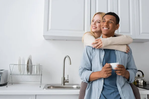 Jovem mulher loira abraçando namorado afro-americano com xícara de café e olhando para longe na cozinha — Fotografia de Stock