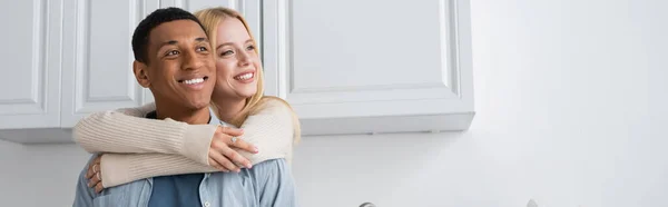 Junge blonde Frau umarmt fröhlichen afrikanisch-amerikanischen Freund und schaut weg in der Küche, Banner — Stockfoto