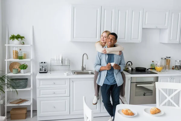 Happy woman sitting on worktop in modern kitchen and embracing african american boyfriend with coffee cup — Stock Photo