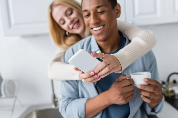 Foyer sélectif du smartphone dans les mains de la femme floue près de l'homme afro-américain avec tasse de café — Photo de stock