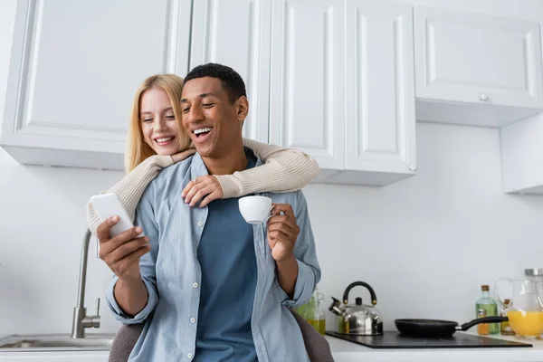 Rindo afro-americano homem segurando xícara de café e smartphone perto de sorrir namorada na cozinha — Fotografia de Stock
