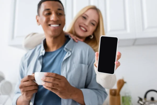 Glücklich blonde Frau hält Smartphone mit leerem Bildschirm in der Nähe afrikanisch-amerikanischer Mann mit Kaffeetasse auf verschwommenem Hintergrund — Stockfoto