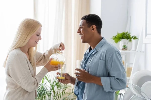 Side view of excited african american man holding glasses near blonde girlfriend pouring fresh orange juice — Stock Photo