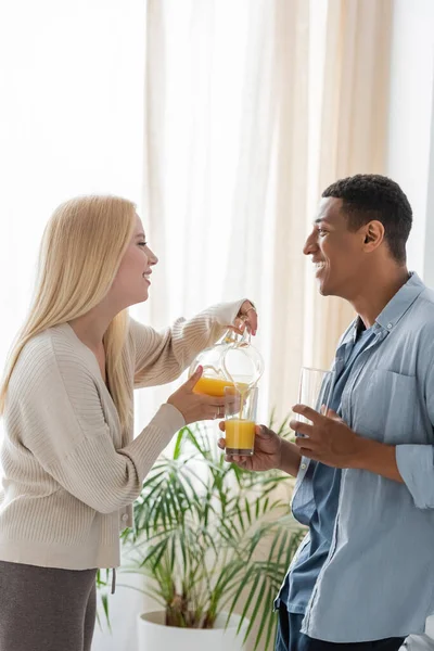 Вид сбоку блондинки, наливающей апельсиновый сок рядом с африканским бойфрендом, держащим очки на кухне — стоковое фото