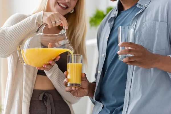 Vista parcial de la mujer sonriente vertiendo jugo de naranja cerca de novio afroamericano con gafas - foto de stock