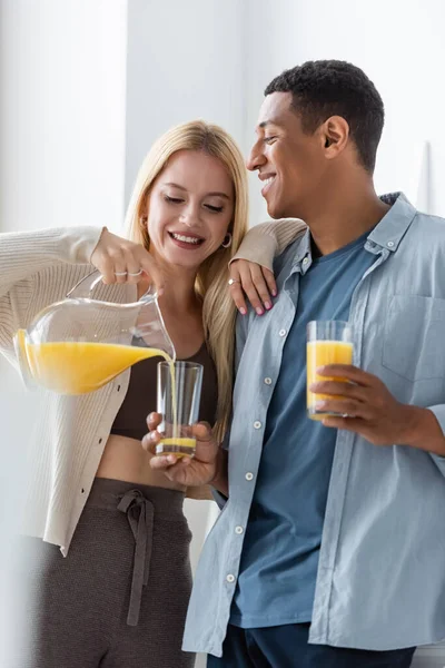 Улыбающаяся блондинка, опирающаяся на африканского американского бойфренда, наливая апельсиновый сок на кухню — стоковое фото