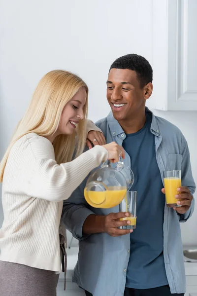 Feliz africano americano hombre celebración gafas cerca joven rubia novia verter naranja jugo - foto de stock