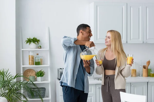 Молодая блондинка в очках улыбается рядом с африканским американским бойфрендом наливая апельсиновый сок на кухню — стоковое фото