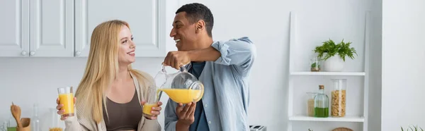 Cheerful african american man looking at blonde girlfriend and pouring fresh orange juice in kitchen, banner — Stock Photo