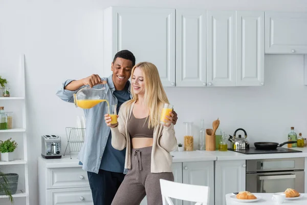 Happy african american man pouring orange juice near pretty girlfriend holding glasses in kitchen — Stock Photo