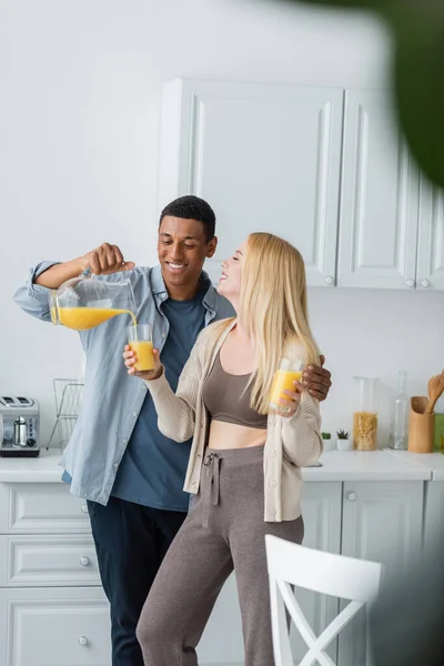 Веселый африканский американец наливает апельсиновый сок рядом с молодой блондинкой, стоящей в очках на кухне — стоковое фото
