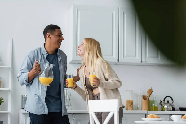 Couple multiethnique excité avec du jus d'orange frais se regardant dans la cuisine sur le premier plan flou — Photo de stock