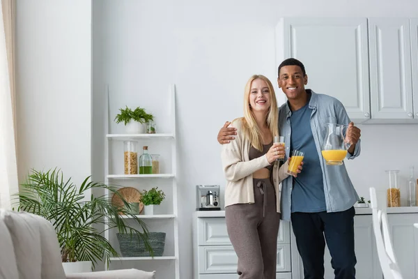 Alegre pareja interracial celebración de jugo de naranja y mirando a la cámara en la cocina moderna - foto de stock