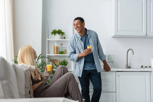 Felice uomo africano americano con un bicchiere di succo d'arancia fresco in piedi vicino alla ragazza seduta sul divano sfocato in cucina — Foto stock