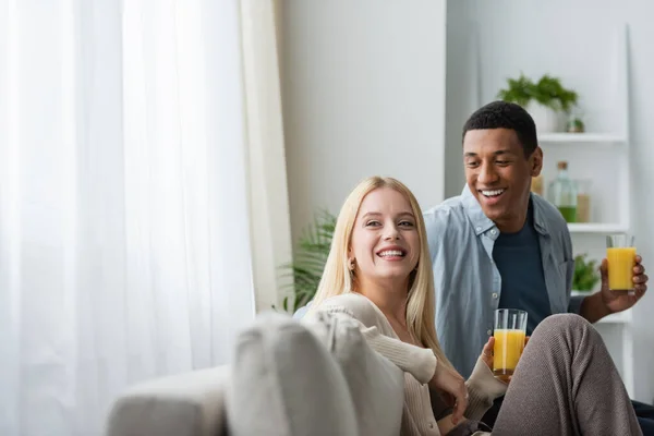 Hombre americano africano alegre y mujer rubia sonriente sentado con jugo de naranja en el sofá en la cocina - foto de stock
