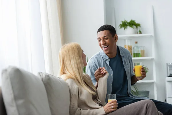 Interracial couple with glasses of orange juice holding hands on couch in kitchen — Stock Photo